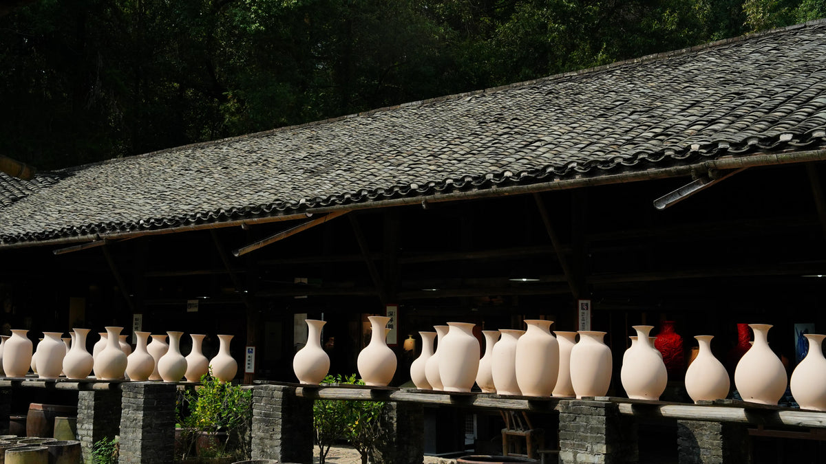 Rows of unglazed Saragaia raw clay ceramic vases drying outdoors on wooden racks under the traditional tiled eaves of a pottery workshop.