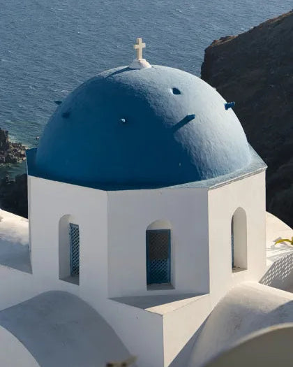 A traditional whitewashed Greek building with a vibrant blue domed roof and cross facing the ocean in Santorini, serving as the Mediterranean aesthetic inspiration for Saragaia's ceramic design.