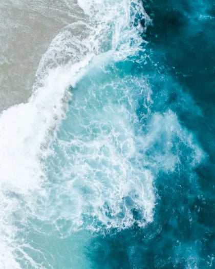 Aerial top-down shot of turquoise ocean water and swirling white froth crashing against the shore, capturing the coastal wave inspiration for Saragaia's ceramic glazes.