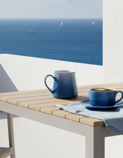 A Saragaia blue gradient coffee cup and mug placed on a outdoor wooden table overlooking a bright blue ocean with white sailboats.