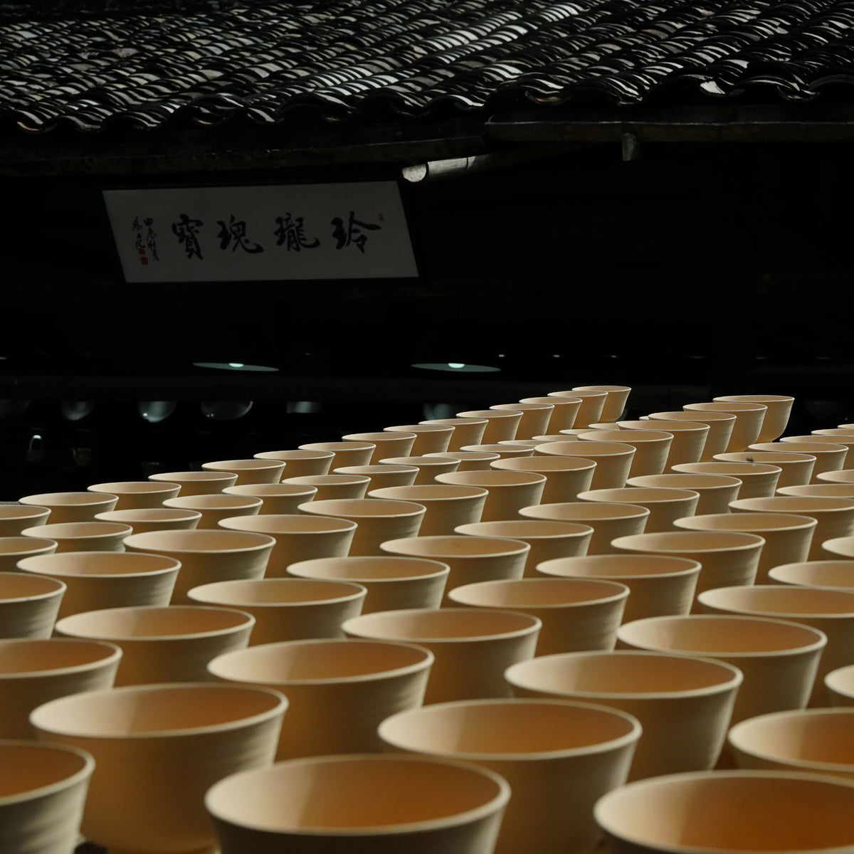Rows of Saragaia ceramic cups in raw clay stage neatly arranged on a table for drying under the traditional tiled roof of an authentic pottery workshop.