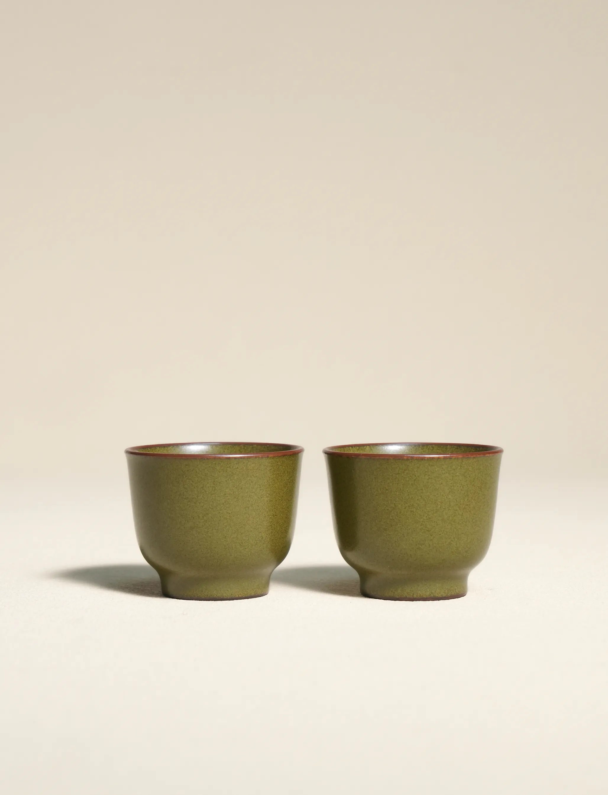 A side-by-side shot of a pair of master-made Saragaia ceramic teacups, finished in a traditional matte green tea-dust glaze with natural brown-accented rims.