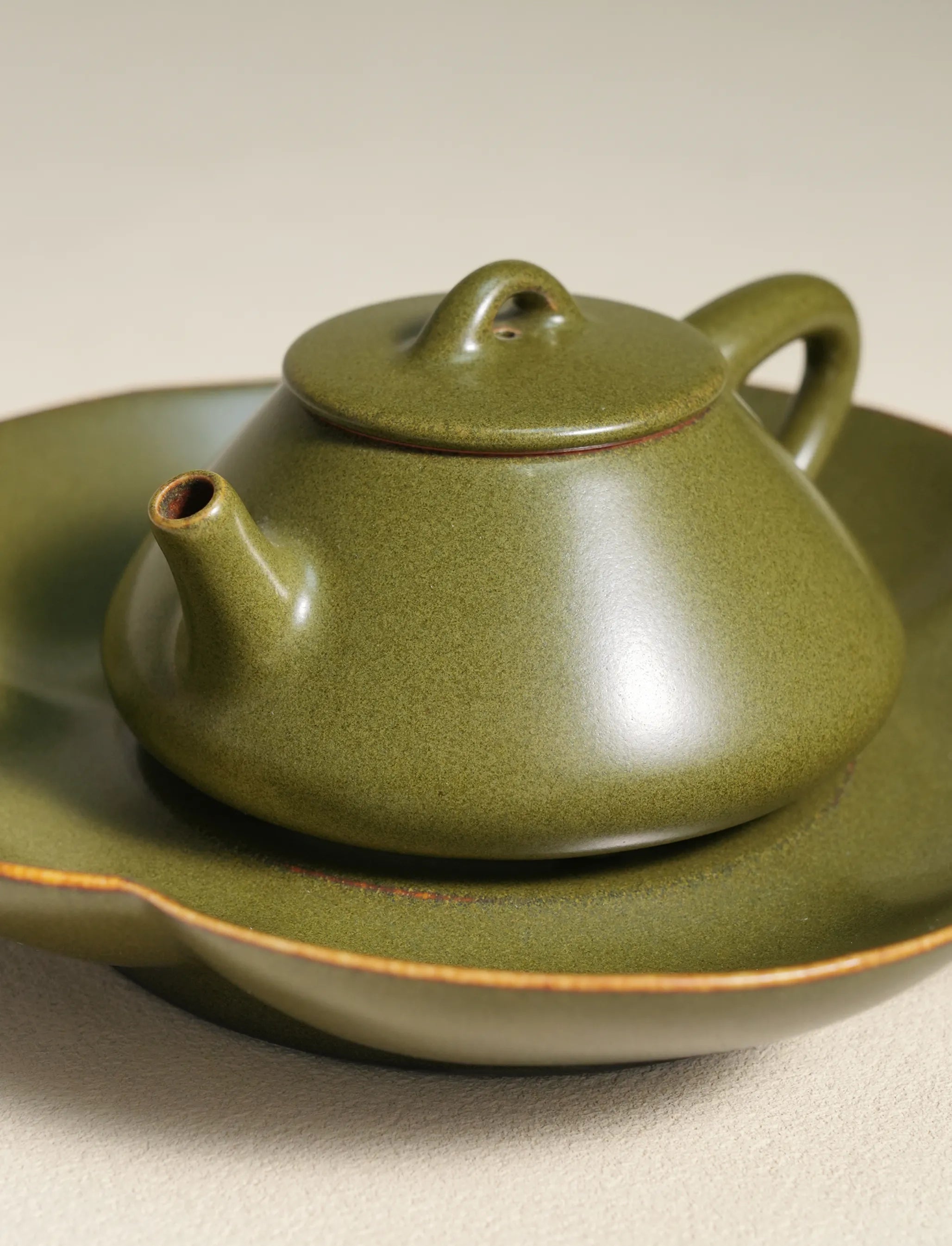 A detailed close-up shot of a master-made Saragaia ceramic teapot resting on a matching tea boat, showcasing the fine texture of the traditional matte green tea-dust glaze and the natural brown edges of the rims.