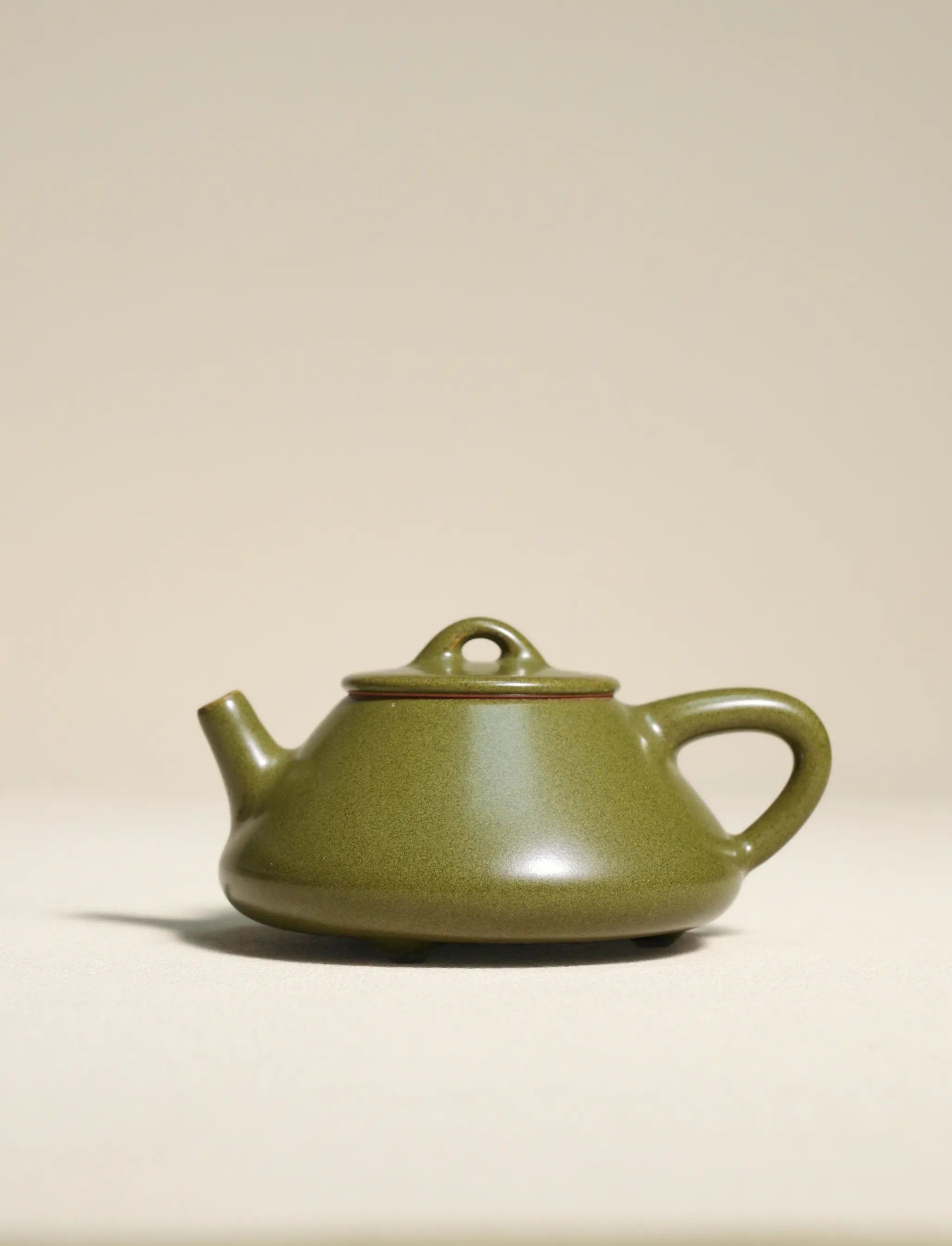 A close-up profile shot of a master-made Saragaia ceramic teapot with small feet, coated in a matte green tea-dust glaze showcasing subtle texture and a natural brown rim accent.