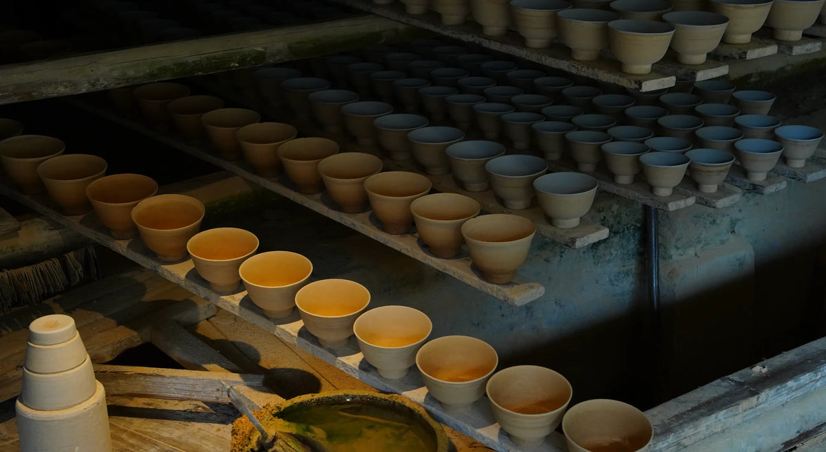 Rows of Saragaia raw ceramic teabowls lined up on long wooden shelves in a quiet drying room prior to glazing and firing.
