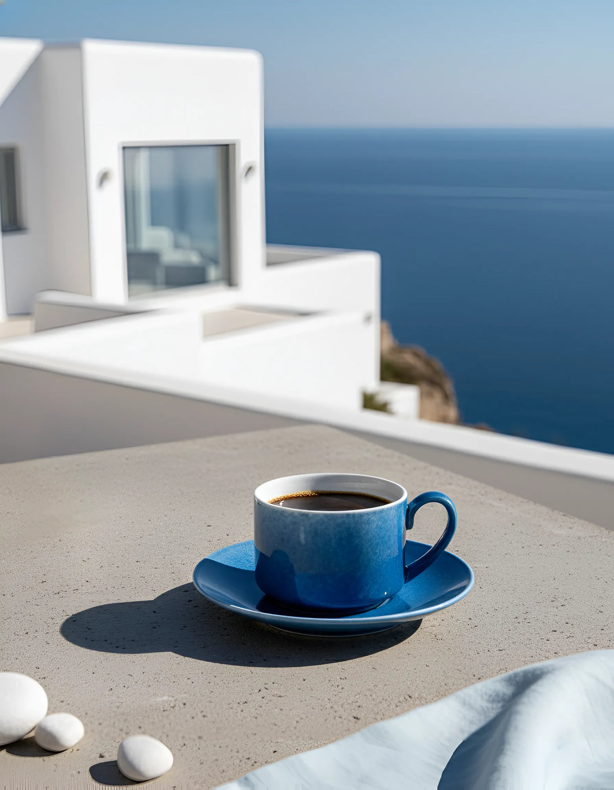 A Saragaia blue gradient ceramic coffee cup and saucer sitting on a concrete tabletop overlooking a Mediterranean white building and the deep blue ocean.