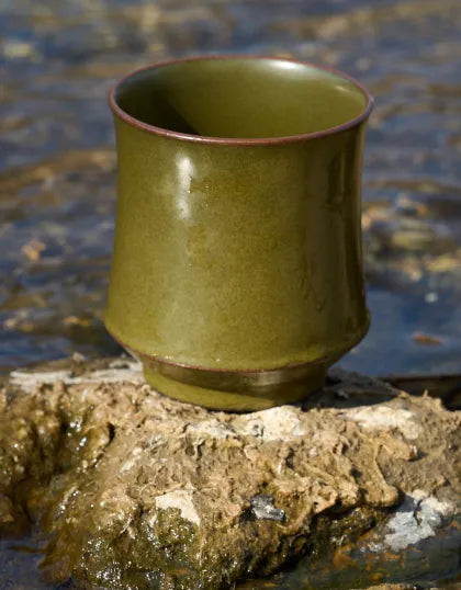 A Saragaia tea-dust glazed ceramic cup resting on a natural mossy stone at the edge of a flowing freshwater stream.