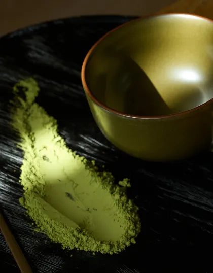 Matcha tea powder dusted on a dark wooden surface next to a Saragaia tea-dust glazed ceramic bowl under direct dramatic lighting.