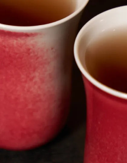 A close-up view of two Saragaia ceramic cups filled with hot tea, showing the soft white-to-red gradient of the peach blossom glaze on their rims.