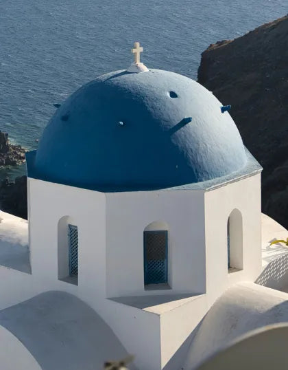 A traditional whitewashed Greek building with a vibrant blue domed roof and cross facing the ocean in Santorini, serving as the Mediterranean aesthetic inspiration for Saragaia's ceramic design.