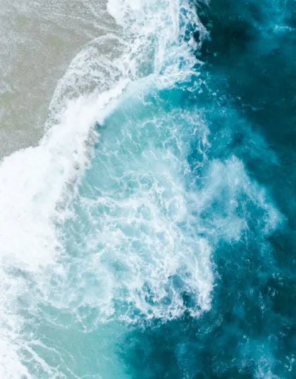 Aerial top-down shot of turquoise ocean water and swirling white froth crashing against the shore, capturing the coastal wave inspiration for Saragaia's ceramic glazes.