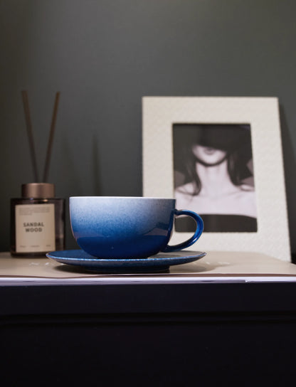 A close-up shot of a master-made Saragaia sprinkling blue ceramic latte cup and saucer set resting on a book, emphasizing its wide, rounded bowl shape and the beautiful gradient of the sprinkling blue glaze
